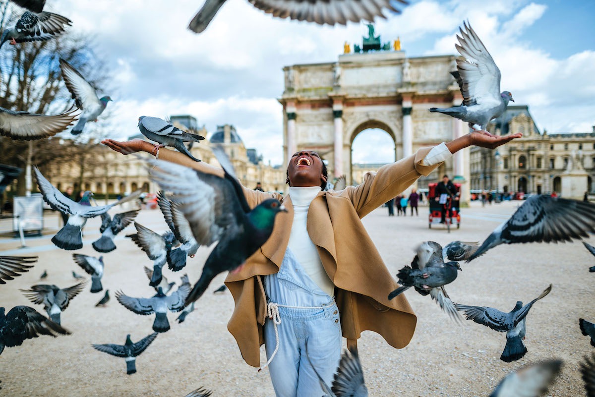 Young smiling woman among doves flying in front of Triumph Arch of Carousel in Paris, France.