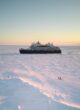 Le Commandant Charcot, Ponant's luxury polar icebreaker, sails through a stark, white landscape of solid ice, dotted with the tiny figures of people walking near the ship.