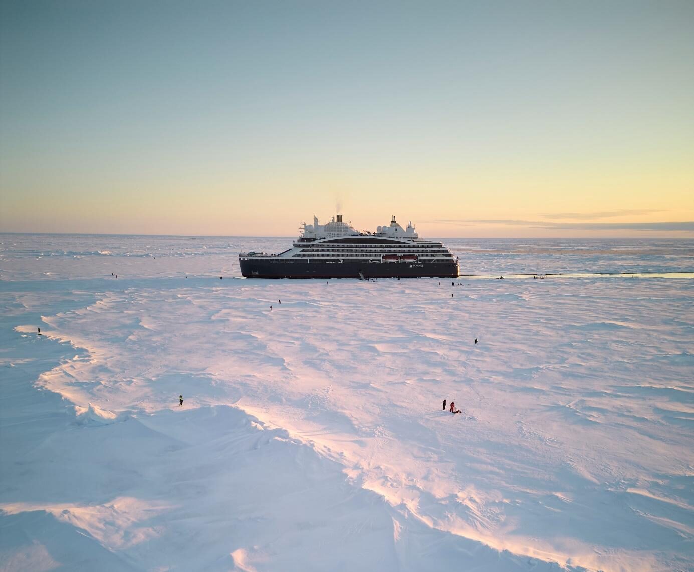 Le Commandant Charcot, Ponant's luxury polar icebreaker, sails through a stark, white landscape of solid ice, dotted with the tiny figures of people walking near the ship.