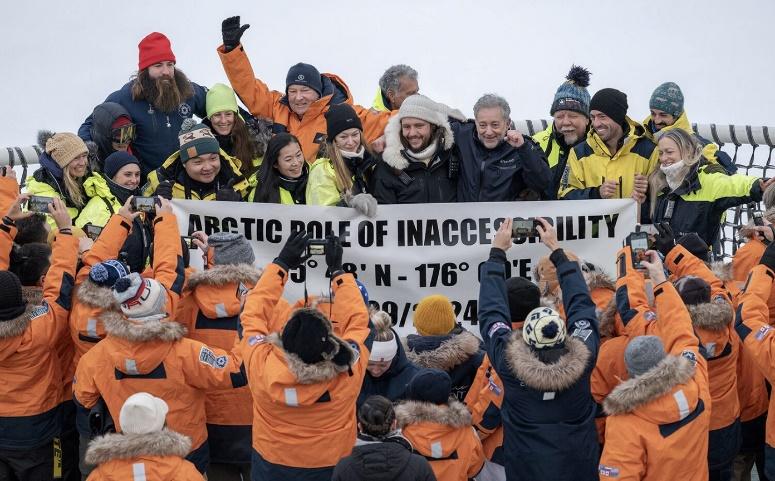 A crowd of travelers in orange parkas holds up a NORTHERN POLE OF INACCESSIBILITY sign on the deck of Ponant's Le Commandant Charcot