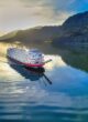 The paddlewheeler American Queen plies a smoothly reflective river in early morning light, a forested mountain rising behind it.