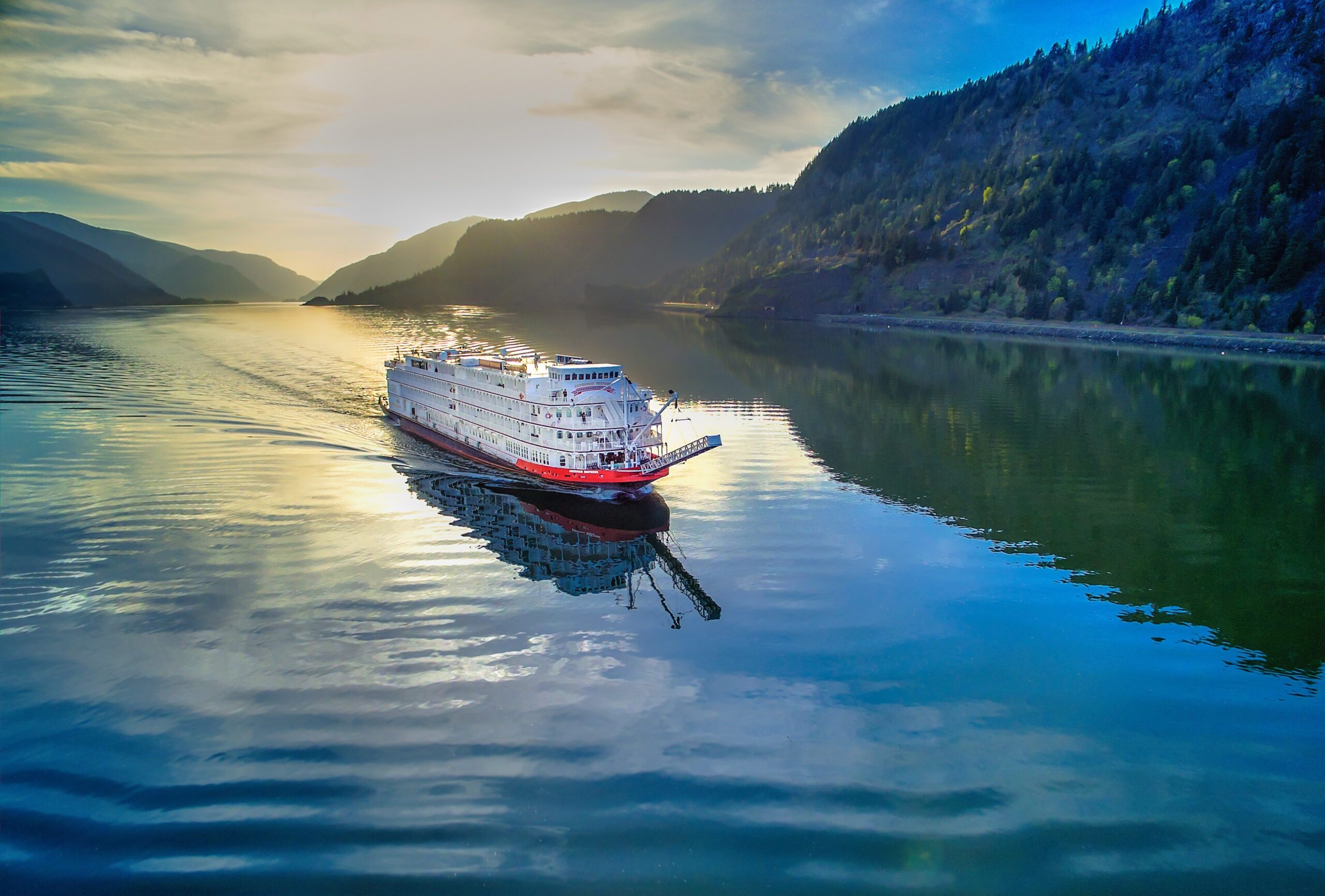 The paddlewheeler American Queen plies a smoothly reflective river in early morning light, a forested mountain rising behind it.