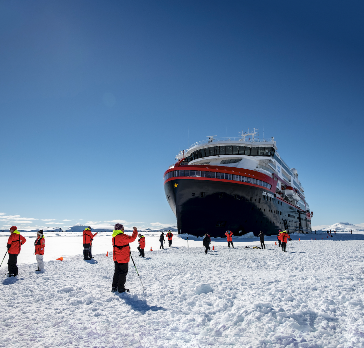 Orange-clad guests and expedition crew stand on the ice surrounding MS Roald Amundsen, polar expedition ship.